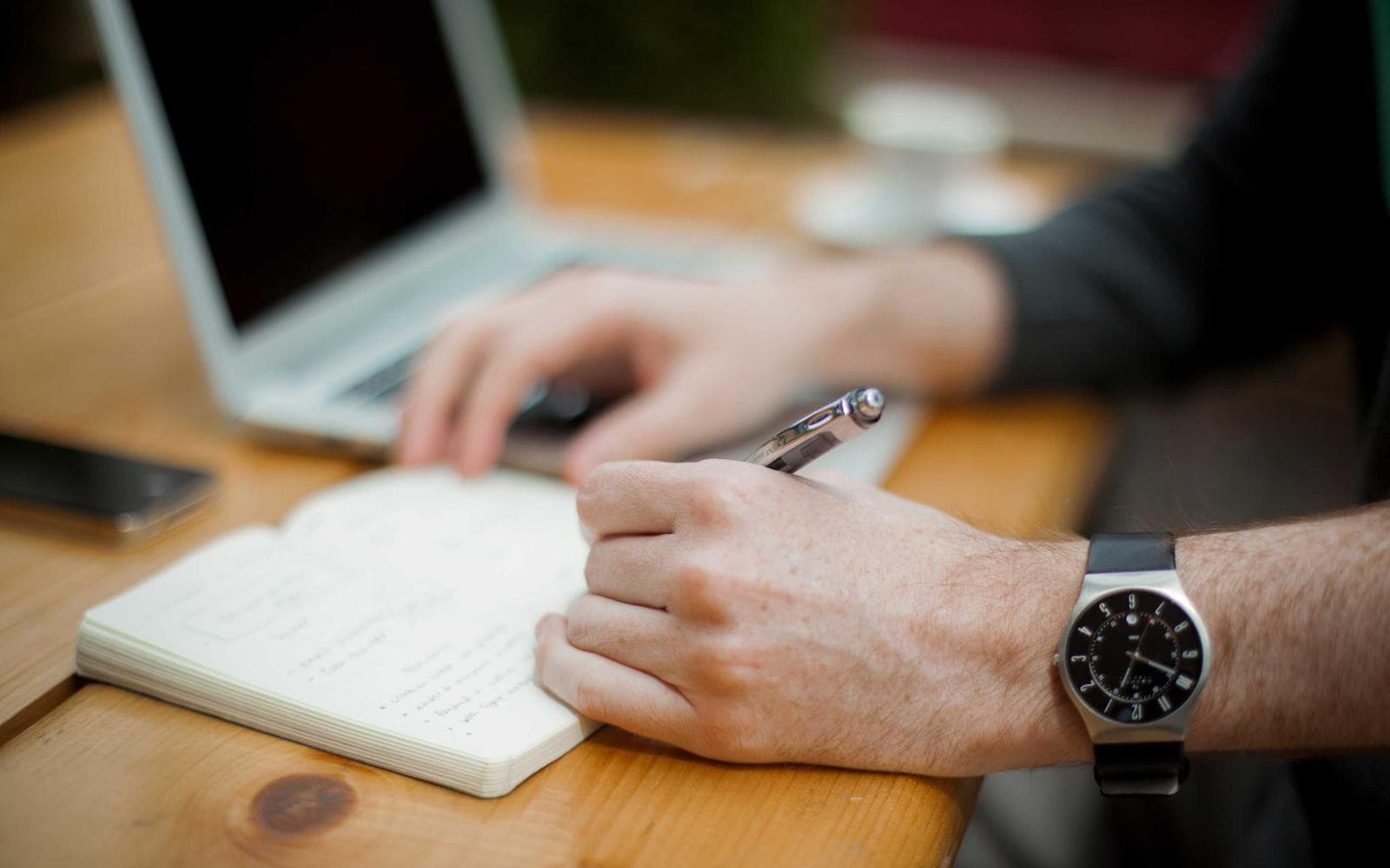 Hands writing in a notebook beside an open laptop.
